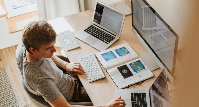 A man multitasks with laptops and a desktop, coding in a home office setting.